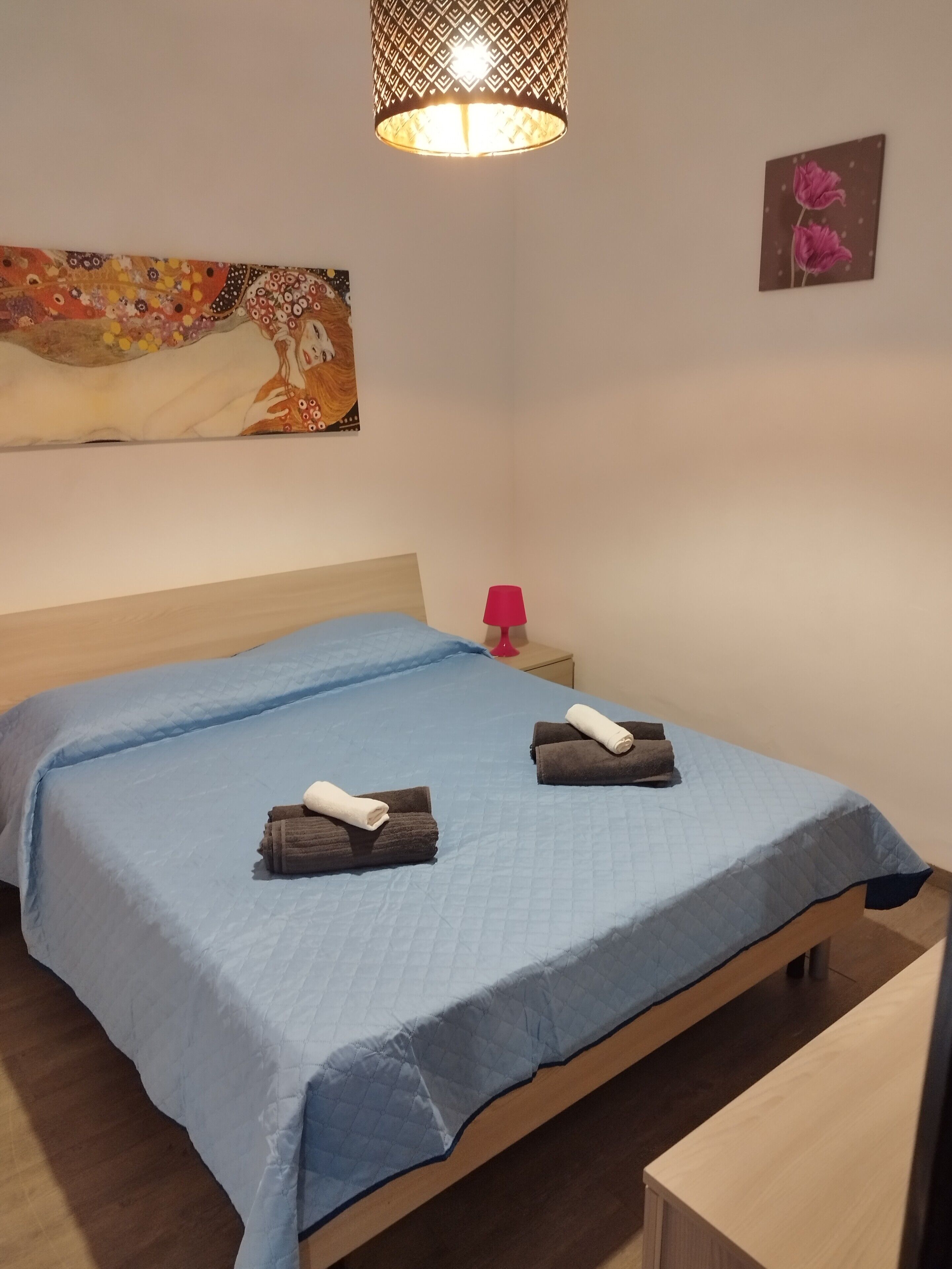 A bedroom with a bed, a bedside table with a lamp, and a painting on the wall.
