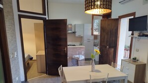 A dining area with a table, chairs, a vase with flowers, a television, and a kitchen in the background.