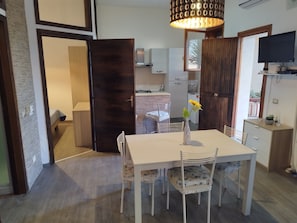 A dining area with a table, chairs, a vase with flowers, a television, and a kitchen in the background.