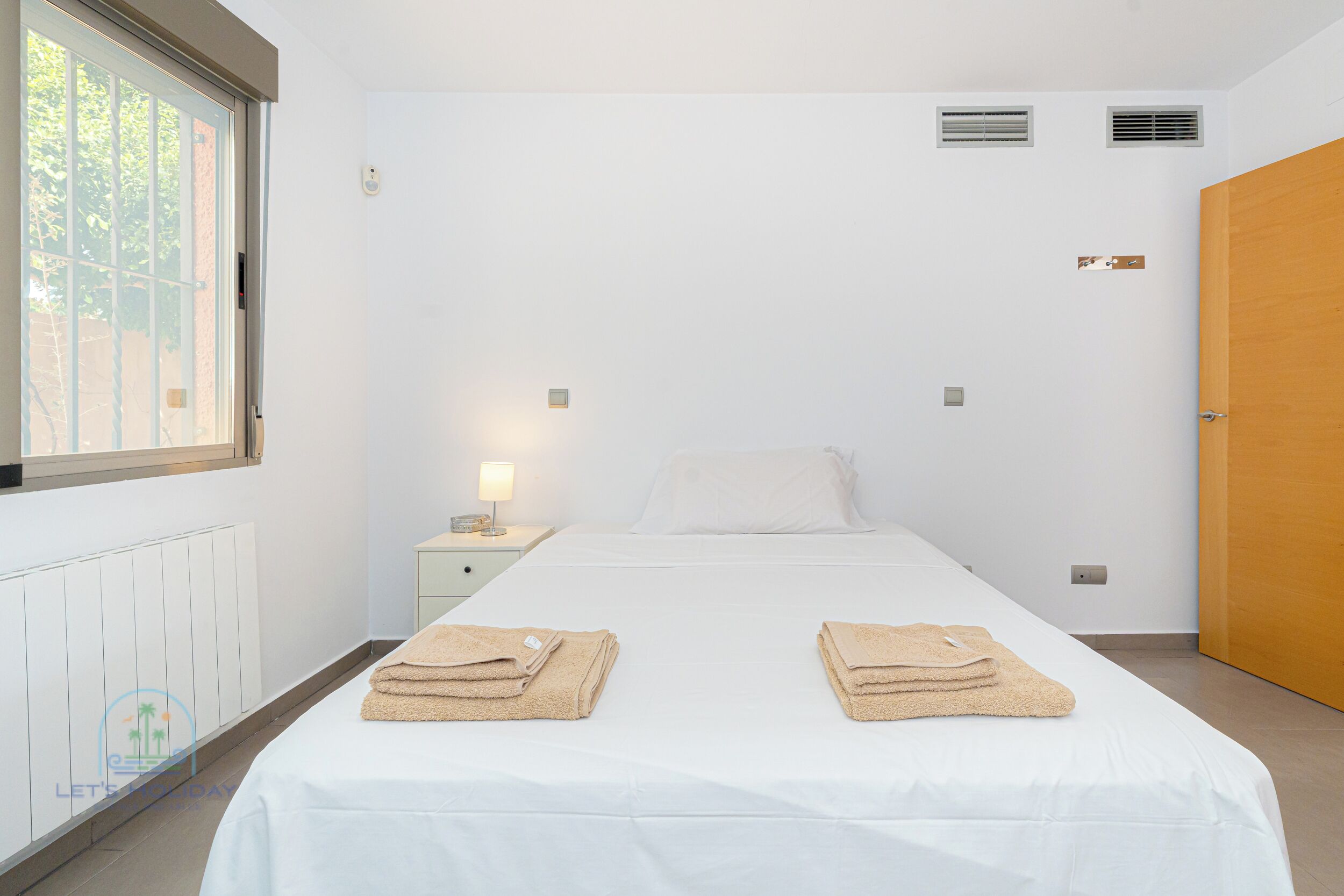 Ein helles, modernes Hotelzimmer mit einem großen Bett, zwei Handtüchern auf dem Bett, einem Nachttisch mit Lampe, einem Fenster und einer Tür.