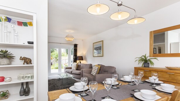 Ein Essbereich mit einem Holztisch, gedecktem für sechs Personen, weißen Tellern und Weingläsern. Im Hintergrund befinden sich eine Couch und ein Schrank.