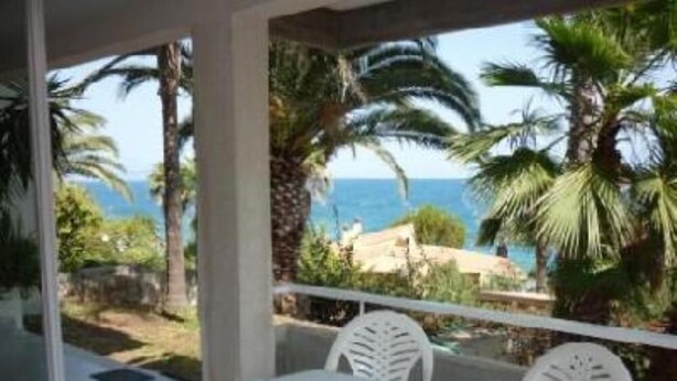A balcony with a white table and chairs, overlooking a coastal area with palm trees and the sea.