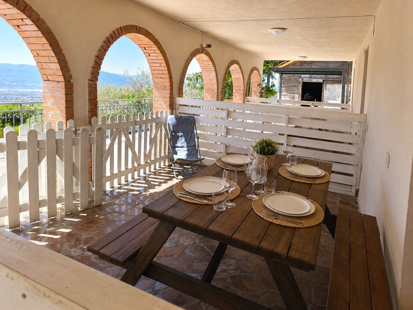 Une terrasse extérieure avec une table en bois dressée pour le repas, des arches en briques, une clôture blanche et une vue sur le paysage.