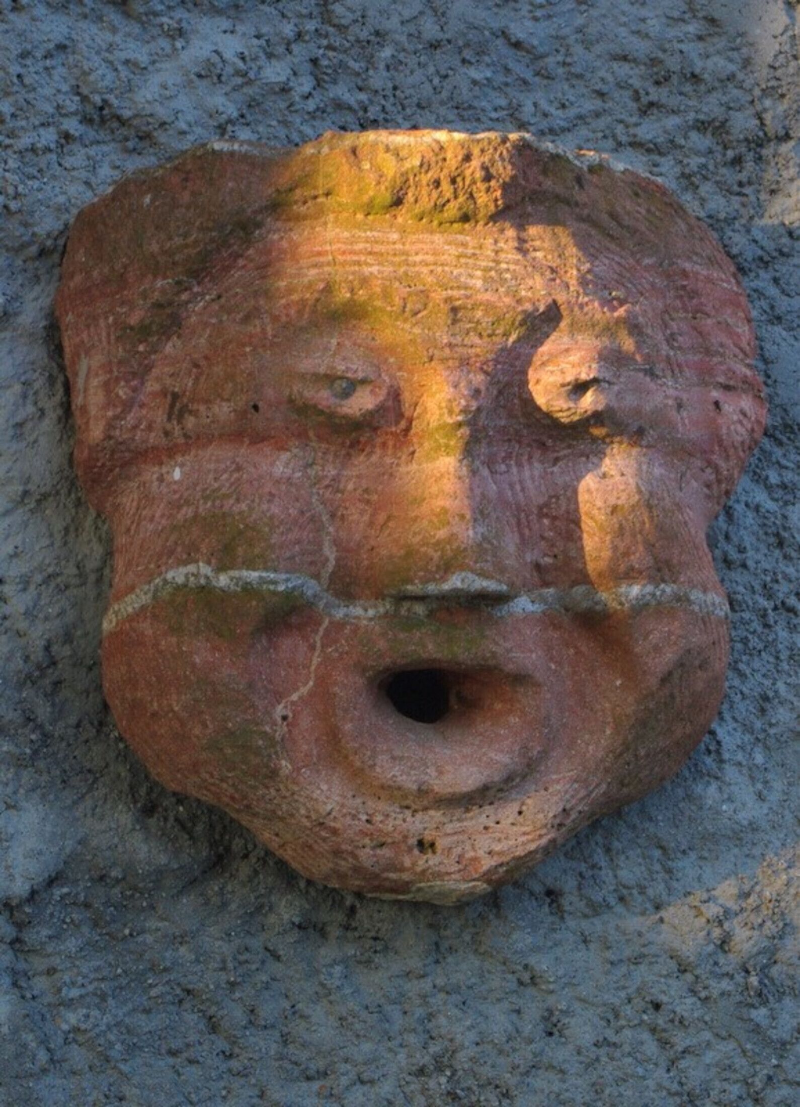 A stone face sculpture with a circular eye-hole and a mouth, set against a textured stone background.