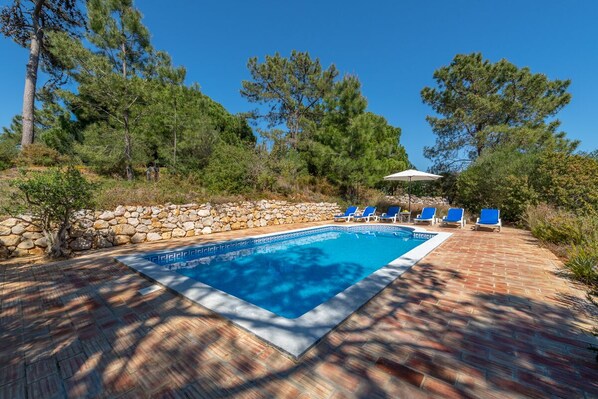 A rectangular swimming pool with blue water, surrounded by a brick patio. There are blue lounge chairs and a white umbrella.