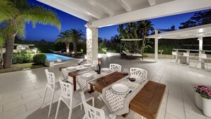 Un área de comedor al aire libre con piscina, muebles blancos y una pérgola.