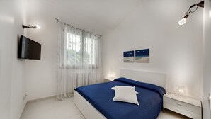 Un dormitorio moderno con una cama, un televisor montado en la pared, una mesita de noche con lámpara y una ventana con cortinas traslúcidas.