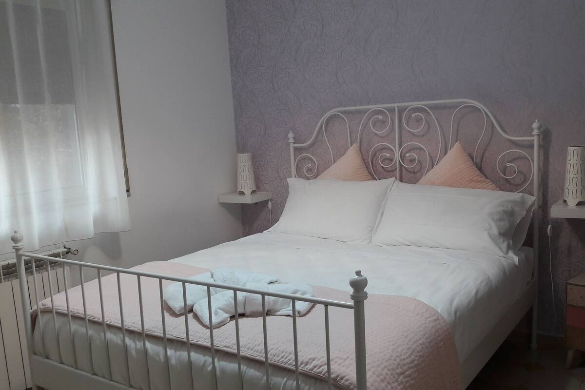 Una cama bien tendida con estructura metálica, ropa de cama blanca y un plaid rosa. Hay dos mesitas de noche con lámparas, una mesita auxiliar y una ventana con cortinas de encaje.