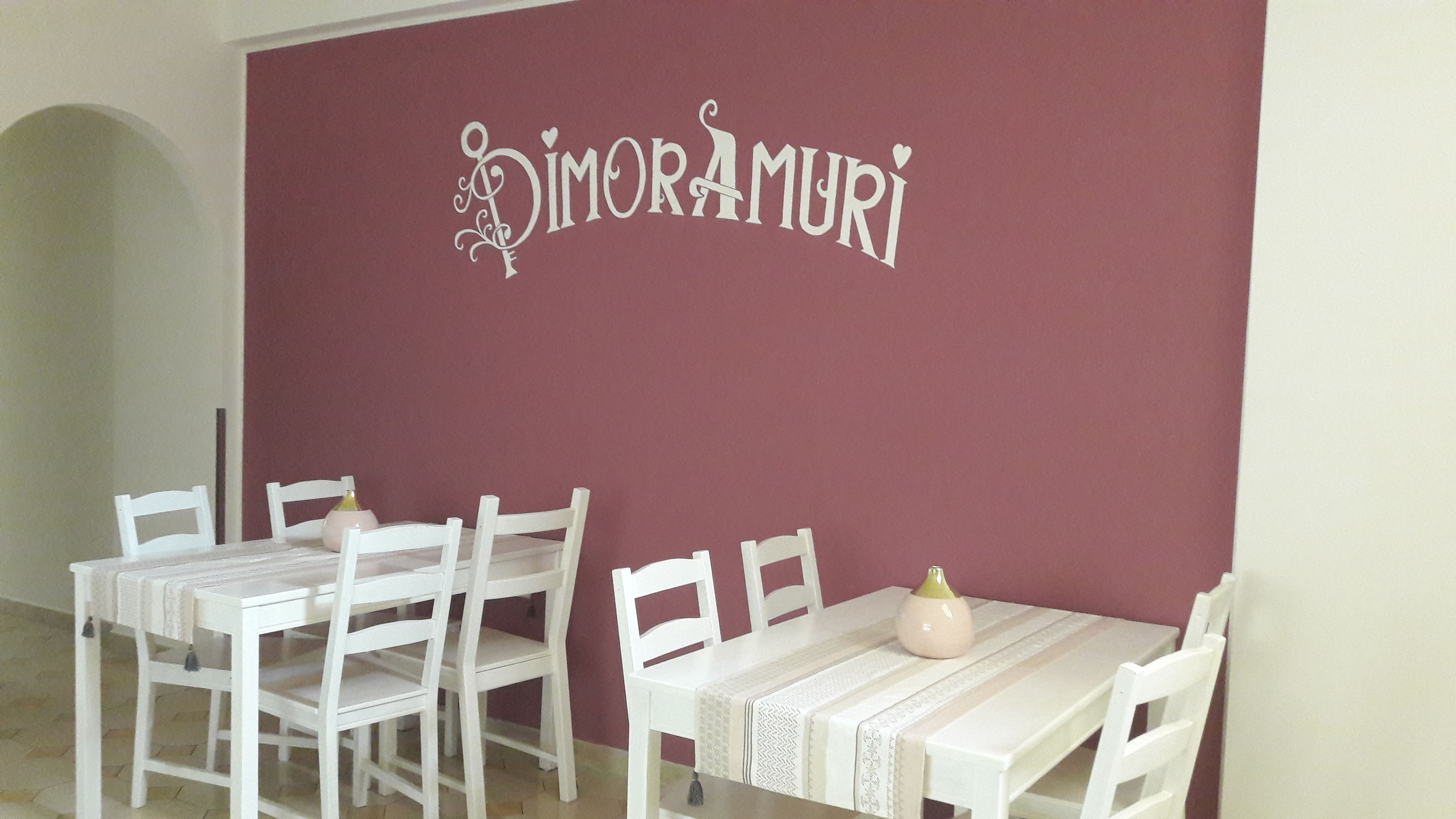 A dining area with white chairs and tables, a maroon wall with the name 'Dimorami' in decorative script, and a small vase on the table.
