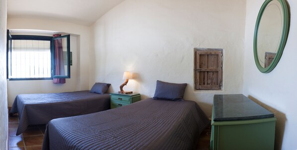 Une chambre avec deux lits, une table de chevet avec une lampe, une petite fenêtre avec des rideaux, un miroir et une armoire en bois.