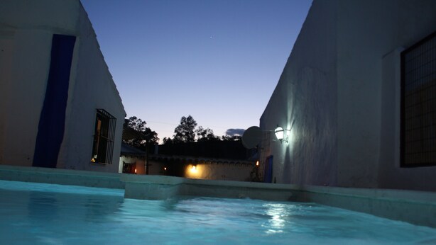 A swimming pool area with two buildings, one with a blue door and another with a light fixture. - River Bed Seat (pruna)