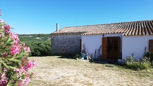 A rustic building with a tiled roof and a stone wall, surrounded by dry grass and flowering plants.