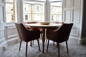 A dining area with a round table and six chairs, large windows with a view of buildings, and a carpeted floor.