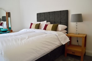 A neatly made bed with white linens, a grey upholstered headboard, and a bedside table with a lamp.