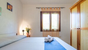 Ein ordentlich bezogenes Bett mit blauem Bettüberzug, ein Holzschrank und ein Fenster mit durchsichtigen Vorhängen.