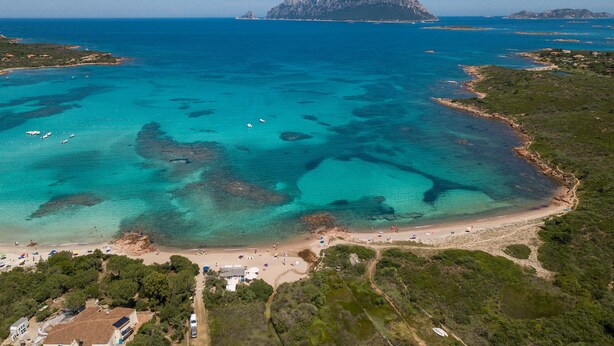 Eine Vogelperspektive auf eine Küstenregion mit klarem türkisblauem Wasser, einem Sandstrand und einer kleinen, felsenigen Insel in der Ferne.