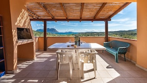 Eine überdachte Terrasse im Freien mit Tisch, Stühlen, einem Grill und Blick auf die Landschaft.