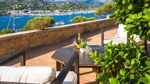 A terrace with wicker furniture, a table with a bottle and glasses, and a view of a marina with boats and a hilly landscape. - VILLA WITH SEA VIEWS, DIRECT ACCESS TO SEA, SWIMMING POOL AND PERFECT LOCATION (Port D'Andratx)