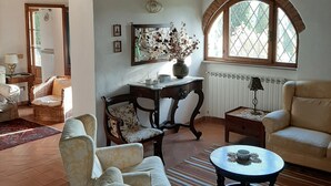 A cozy living room with a high ceiling, wooden beams, a large arched window, and a variety of seating including a sofa, armchairs, and a small table. - Villa Banditello, secluded, panoramic view over Montepulciano valley. (torrita di siena, fraz. montefollonico)