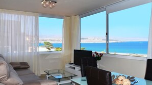 Una sala de estar moderna con un sofá, una mesa de vidrio y una televisión. Hay un ventanal con vistas al mar y a edificios.