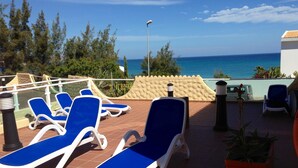 Una terraza con sillones azules, suelo de baldosas, planta en maceta y vista al mar.