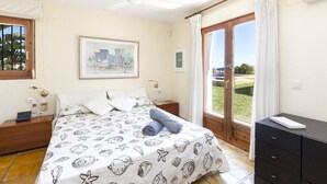 Une chambre à coucher comprenant un lit, une table de chevet, une commode, un ventilateur de plafond, une fenêtre avec des rideaux et un tableau encadré accroché au mur.