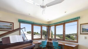 Une pièce avec un piano, un bureau, deux chaises et de grandes fenêtres donnant sur un beau paysage.