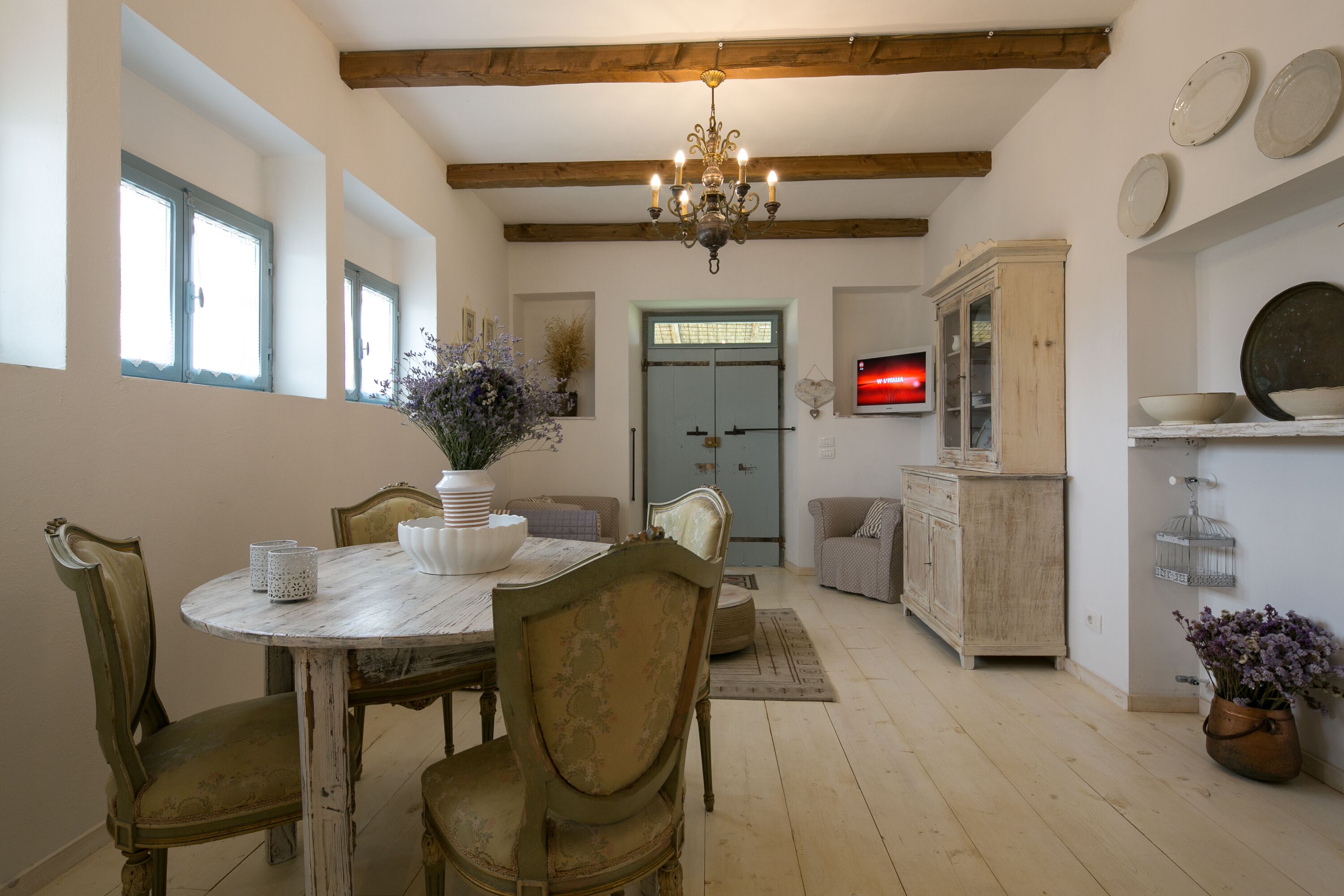 A dining area with a round table, chairs, and a chandelier. There is a cabinet and a television on the wall.