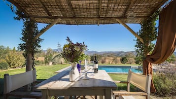 An outdoor seating area with a table, chairs, a vase with flowers, a wine bottle, and glasses. There is a pool in the background.