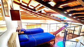 Ein Zimmer mit schrägen Deckenbalken, einem Bett mit blauer Bettdecke, einem Heizkörper und einem Fenster mit Blick.
