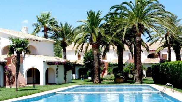 A swimming pool surrounded by palm trees and a building with arches. - Cabins 59 by homeprive (Denia)