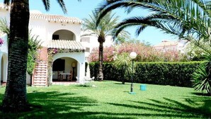 A well-maintained garden with a white building featuring an arched entrance, palm trees, and a neatly trimmed hedge. - Cabins 59 by homeprive (Denia)