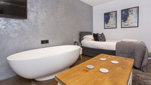 A modern bathroom with a freestanding bathtub, a wooden table with decorative items, a bed with a gray headboard, and two abstract paintings on the wall. - Converted stable room 2 (York)
