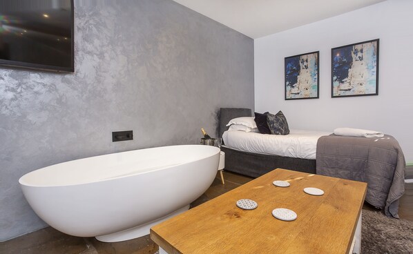 A modern bathroom with a freestanding bathtub, a wooden table with decorative items, a bed with a gray headboard, and two abstract paintings on the wall.