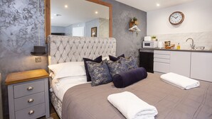 A bedroom with a large bed, a bedside table, a nightstand, a mirror, a wall clock, and a kitchenette with a microwave, sink, and cabinets. - Converted stable room 2 (York)