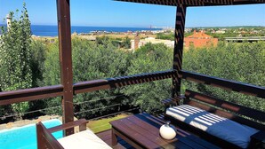 A wooden deck with a pool, lounge chairs, and a view of the sea and cityscape.