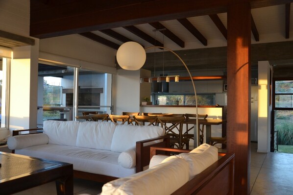 A modern living room with a white sofa, wooden chairs, a dining table, and a kitchen in the background.