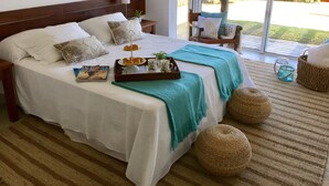 A neatly made bed with a tray of pastries and a book, a large window with a view of greenery, and a cozy chair by the window.