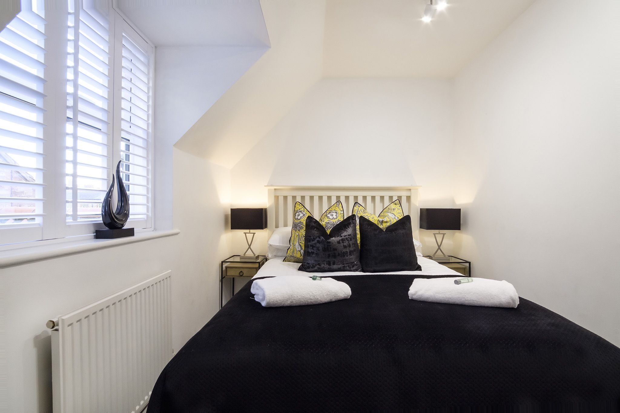 A bedroom with a bed, two bedside tables, a radiator, and a window with blinds.