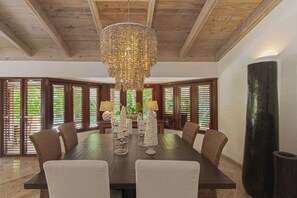 A dining area with a large wooden table, six chairs, a chandelier, and large windows with blinds. - PIÑAS 13 Tropical Holidays in Casa de Campo - 4BDR with private pool and jacuzzi (Casa de Campo)
