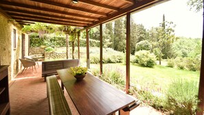 Un portico coperto con tavolo e sedie in legno, un muro in pietra e vista su un giardino rigoglioso.