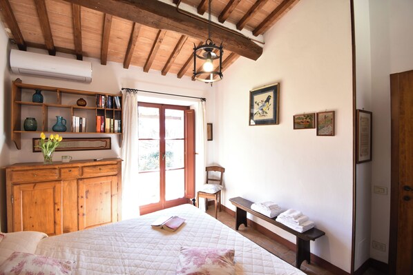 Ein Schlafzimmer mit einem Bett, einer hölzernen Kommode, einem Sessel, einer Bank und einem Regal mit Büchern und Dekoration.