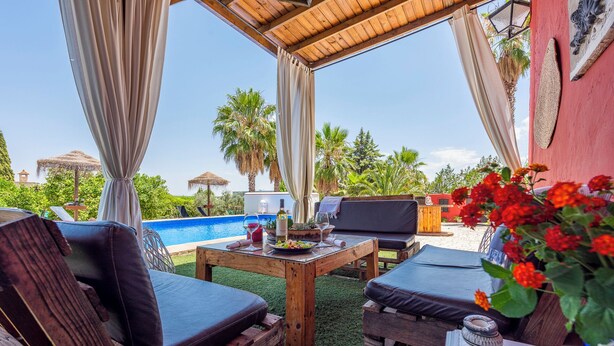 A poolside area with wooden furniture, a table with a bottle and glass, and a view of the pool and greenery. - Villa Felisa Holiday Home, Seville Countryside. (La Puebla de Cazalla)