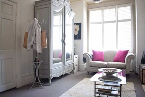 Ein Zimmer mit einem weißen Kleiderschrank, einem weißen Sofa mit rosa Kissen, einem Tisch mit einer Schale und einem Fenster mit Jalousien.