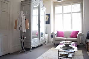 Ein Zimmer mit einem weißen Kleiderschrank, einem weißen Sofa mit rosafarbenen Kissen, einem kleinen Tisch mit einer Schale und einem Fenster mit Jalousien.