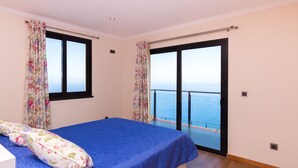 Un dormitorio con cama, cortinas florales, vista al mar a través de una puerta corrediza y un balcón.