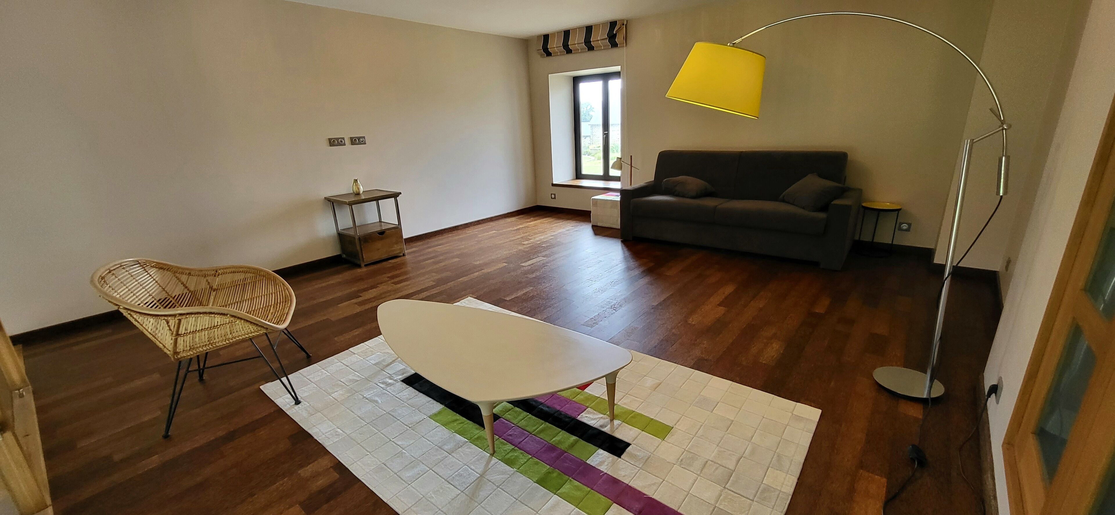Une pièce avec un plancher en bois, une table blanche, une chaise en osier, un canapé gris, un lampadaire jaune et une fenêtre avec des rideaux.