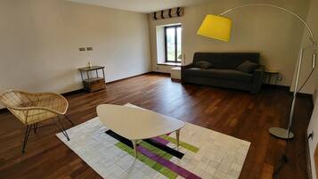 Une pièce avec un plancher en bois, une table blanche, une chaise en osier, un canapé gris, un lampadaire jaune et une fenêtre avec des rideaux.