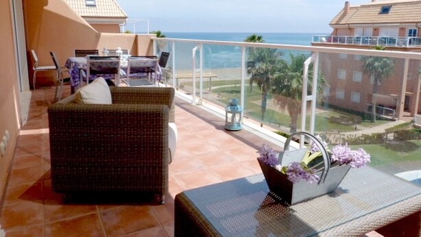 Un balcon agrémenté de meubles en osier, une table avec un vase de fleurs et une vue sur la mer et les bâtiments.