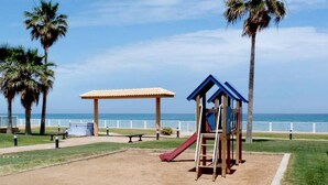 Un terrain de jeux avec un toboggan et une structure d’escalade, entouré de palmiers et offrant une vue sur la plage.